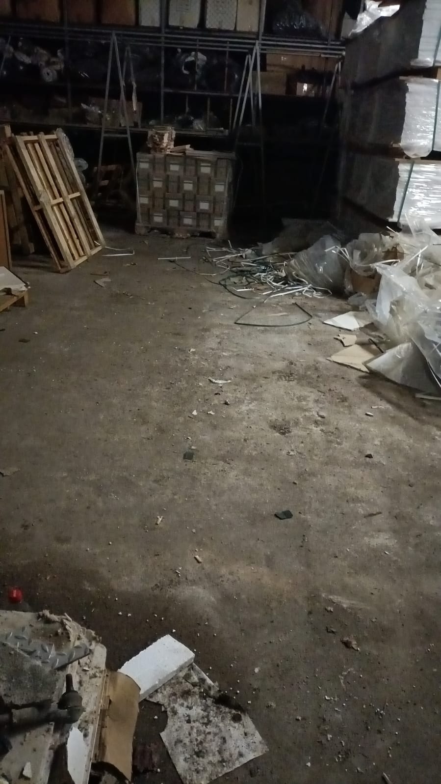 Zaniedbane wnętrze magazynu z porozrzucanymi odpadami, paletami i stertami zapakowanych towarów. Słabe oświetlenie podkreśla stan pomieszczenia.