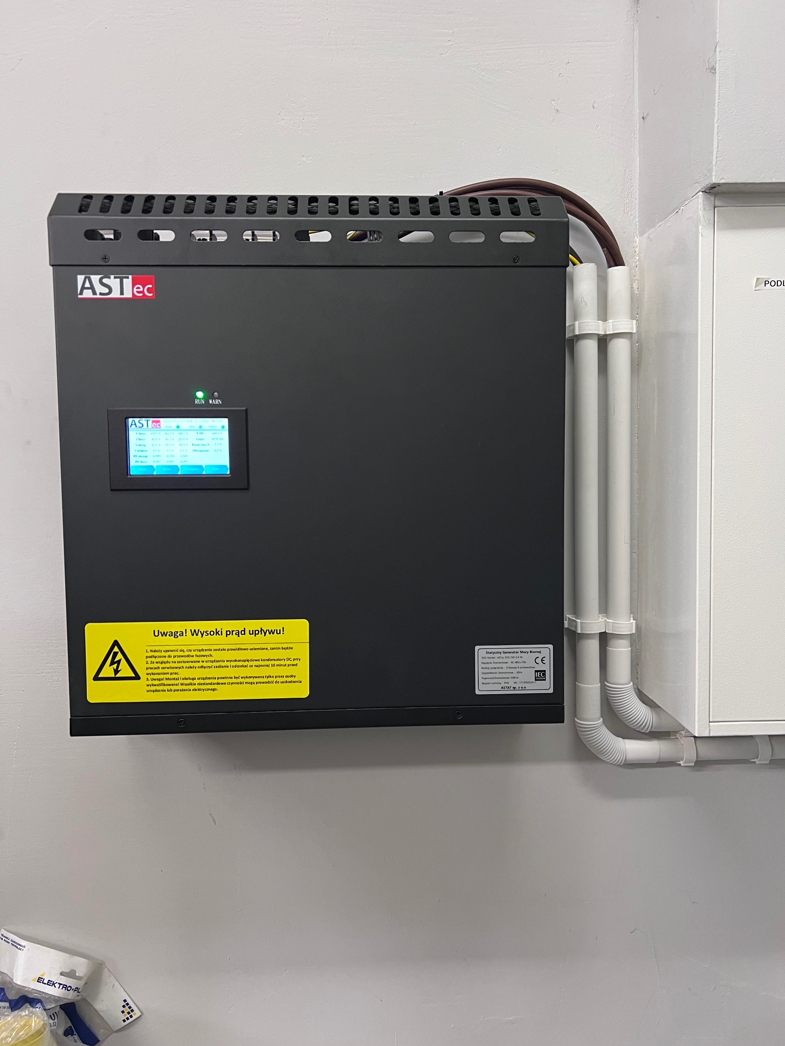 Czarna skrzynka ASTec z ekranem i ostrzeżeniem o wysokim napięciu, zamontowana na ścianie, z widocznymi przewodami i rurami. Element instalacji elektrycznej.