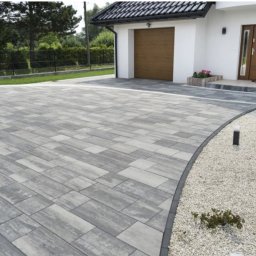 Solidus - Nowoczesny podjazd z szarej kostki brukowej ułożonej we wzór prostokątów, obok biały żwir i niskie krzewy, w tle biały dom z brązowymi drzwiami i bramą garażową.