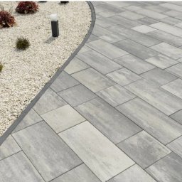 Solidus - Ułożona szara kostka brukowa tworząca ścieżkę w ogrodzie, obok rabata z białym żwirem i roślinami. Widoczne oświetlenie ogrodowe.