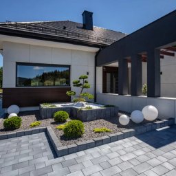 Solidus - Nowoczesny dom jednorodzinny z elewacją w jasnych kolorach, ciemnym dachem i minimalistycznym ogrodem z kostki brukowej oraz dekoracyjnymi kulami.