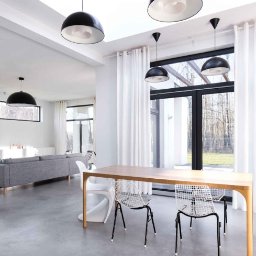 Solidus - Jasne, nowoczesne wnętrze z betonową podłogą, drewnianym stołem i czarnymi lampami wiszącymi. Duże okna z białymi zasłonami wpuszczają naturalne światło.