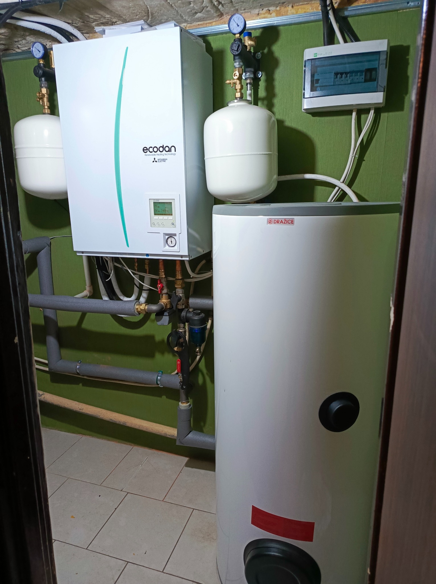 Zestaw urządzeń: pompa ciepła Mitsubishi Ecodan, zbiornik Drazice, instalacja hydrauliczna i elektryczna na tle zielonej ściany w pomieszczeniu technicznym.