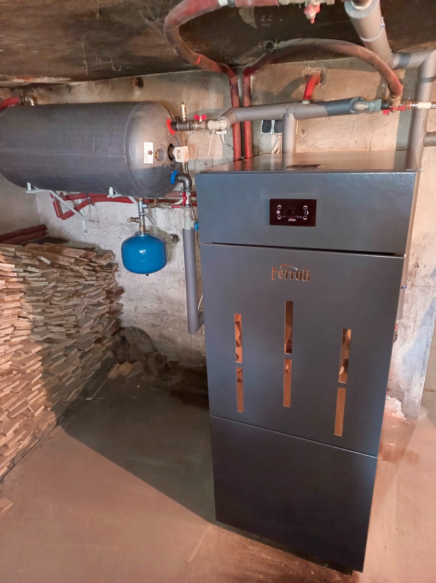 Szary piec grzewczy Ferroli z zbiornikiem akumulacyjnym i instalacją w piwnicy w Słupcy. Widoczne rury i elementy instalacji.