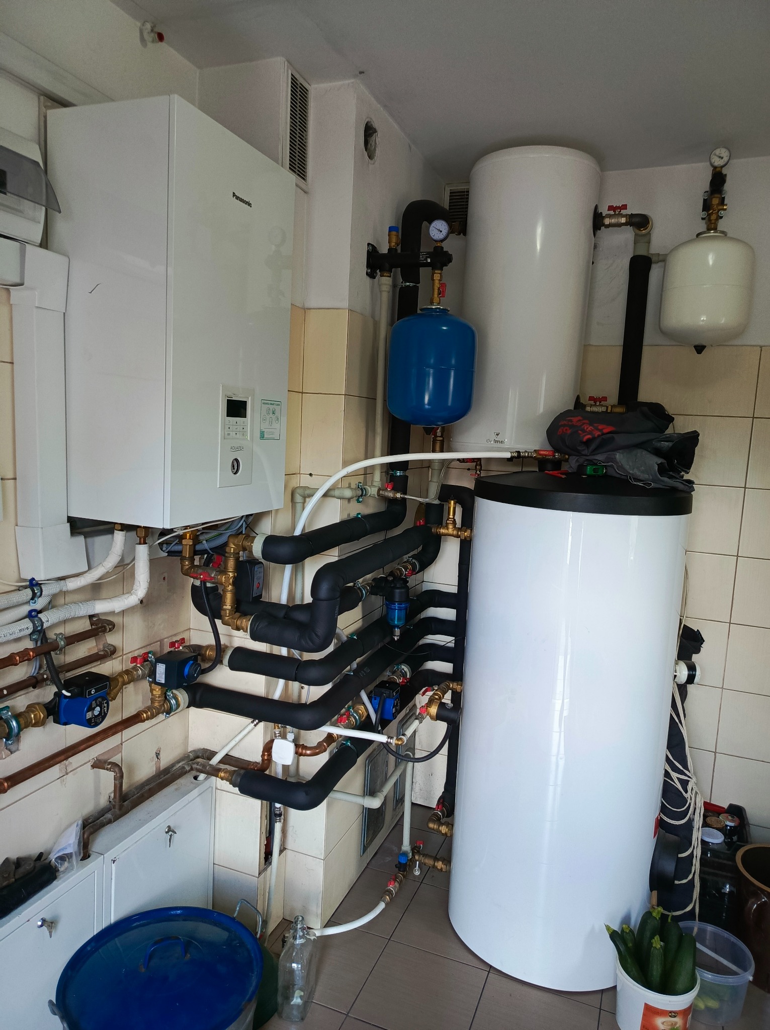 Złożona instalacja z białym zasobnikiem ciepłej wody, kotłem gazowym Panasonic i rurami w pomieszczeniu z wykafelkowanymi ścianami. Widoczne liczne zawory i manometry.