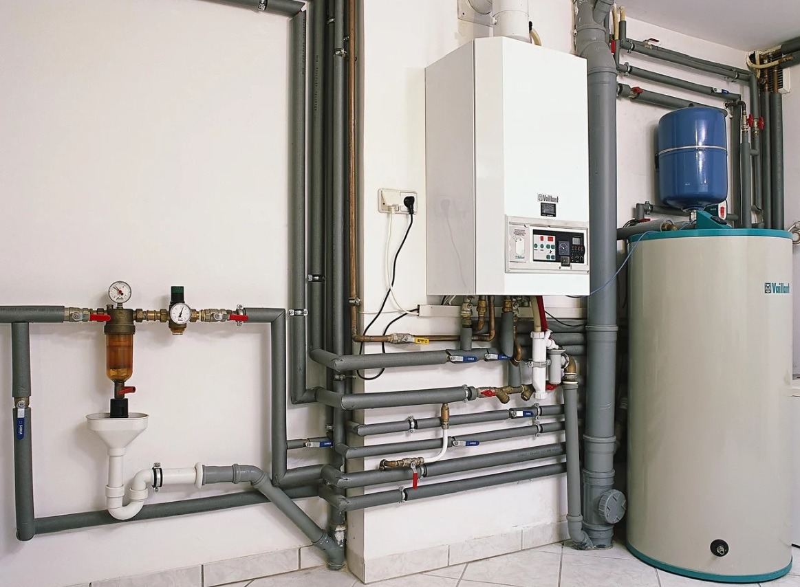 Biały piec gazowy Vaillant z szarymi rurami instalacji CO i wodnej na tle białej ściany i podłogi z płytek. Widoczny zbiornik wyrównawczy i filtr z manometrami.