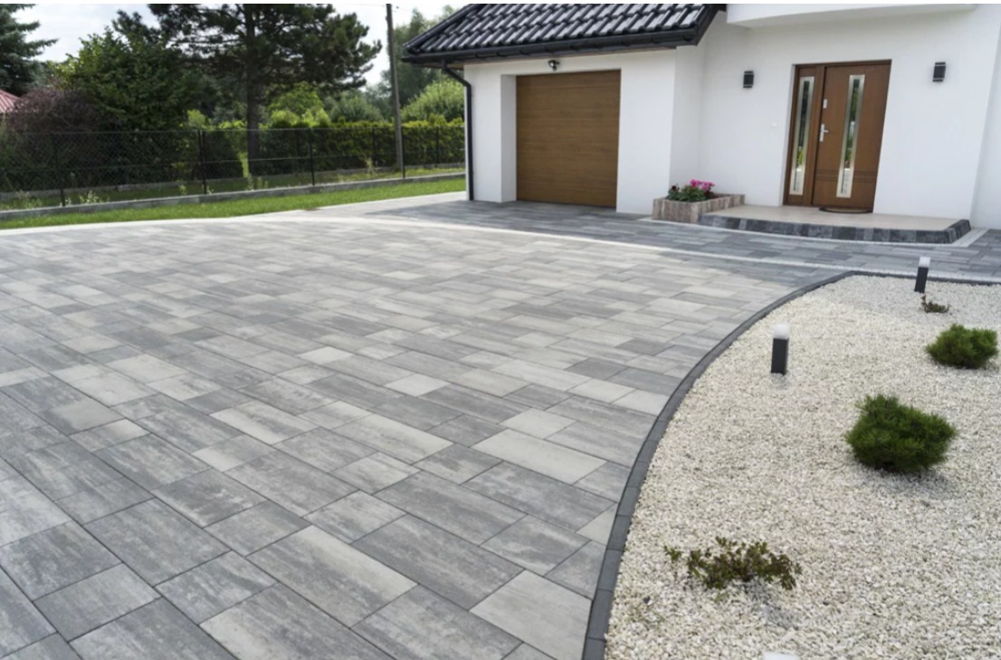 Nowoczesny podjazd z szarej kostki brukowej ułożonej we wzór prostokątów, obok biały żwir i niskie krzewy, w tle biały dom z brązowymi drzwiami i bramą garażową.
