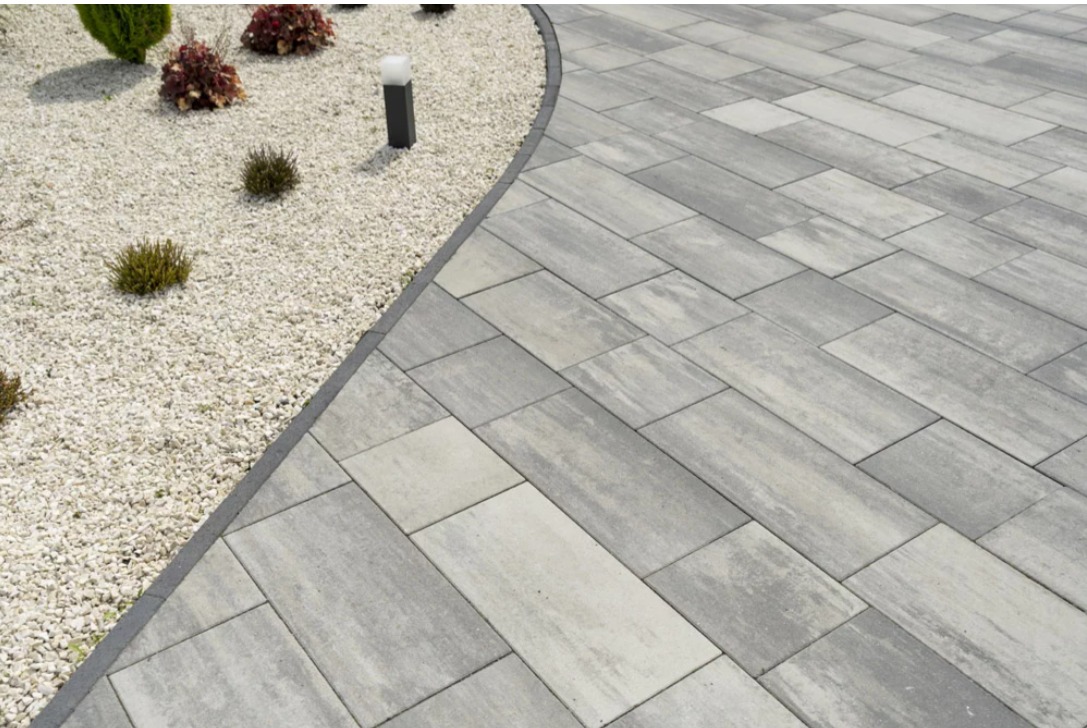 Ułożona szara kostka brukowa tworząca ścieżkę w ogrodzie, obok rabata z białym żwirem i roślinami. Widoczne oświetlenie ogrodowe.