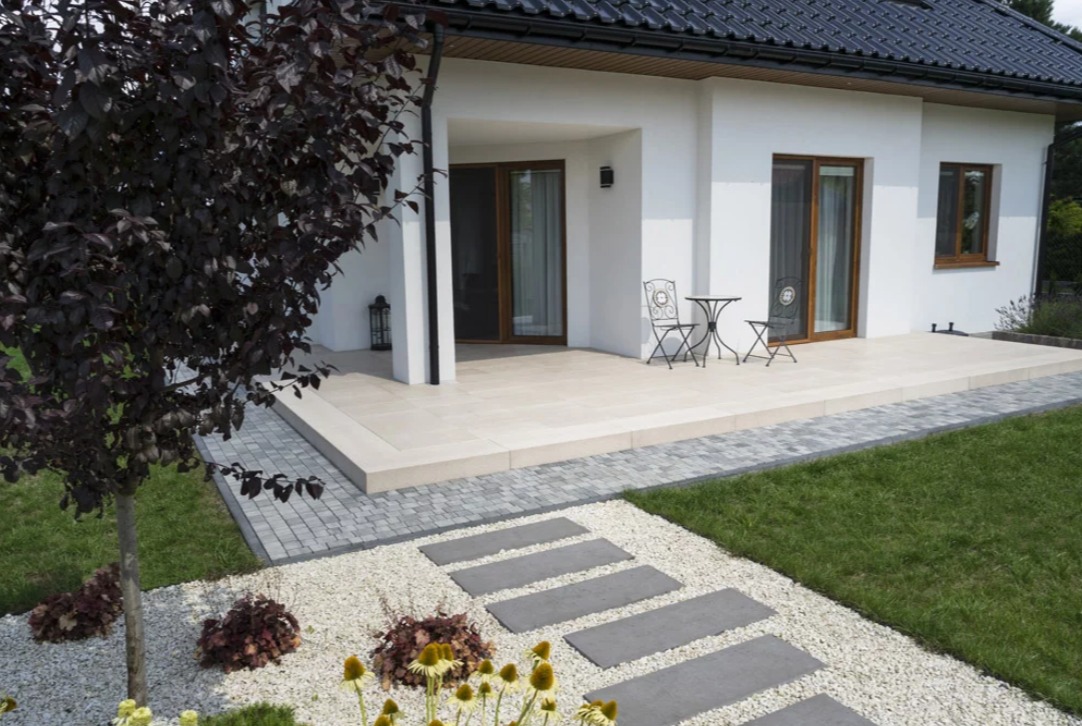 Nowoczesny dom z tarasem wyłożonym jasnymi płytami, stolik i krzesła na zewnątrz. Ścieżka z płyt prowadzi przez biały żwir i zielony trawnik.