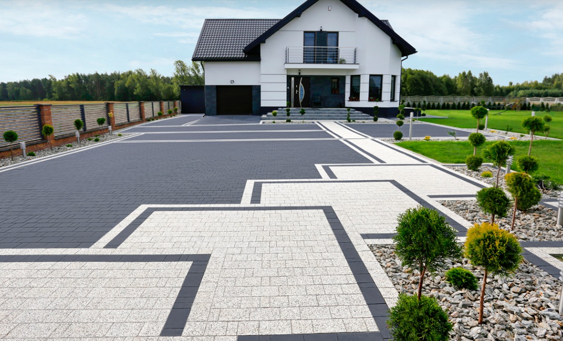 Nowoczesny dom z eleganckim podjazdem z kostki brukowej w dwóch odcieniach szarości, otoczony zielenią i ozdobnymi krzewami. Widok na zadbany ogród i ogrodzenie.