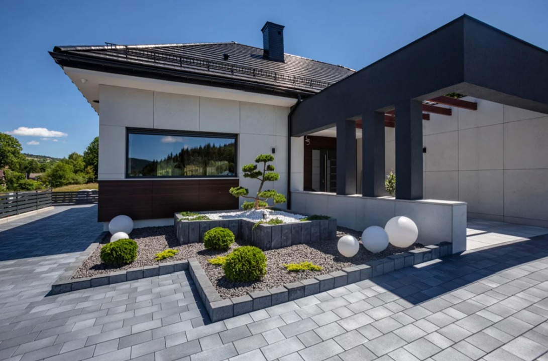 Nowoczesny dom jednorodzinny z elewacją w jasnych kolorach, ciemnym dachem i minimalistycznym ogrodem z kostki brukowej oraz dekoracyjnymi kulami.