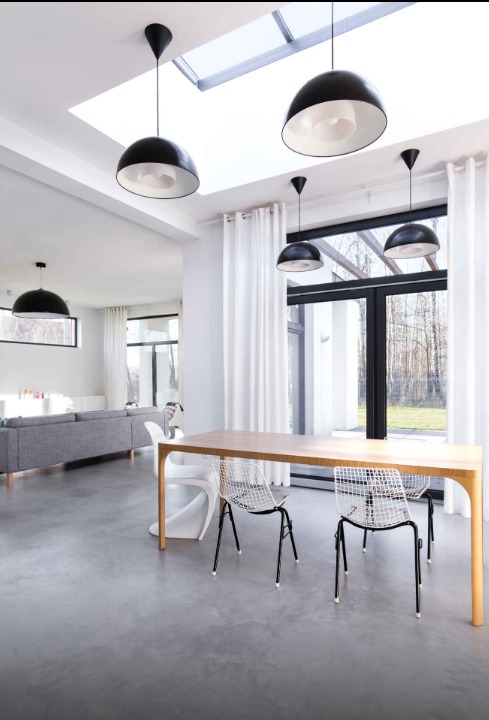 Jasne, nowoczesne wnętrze z betonową podłogą, drewnianym stołem i czarnymi lampami wiszącymi. Duże okna z białymi zasłonami wpuszczają naturalne światło.
