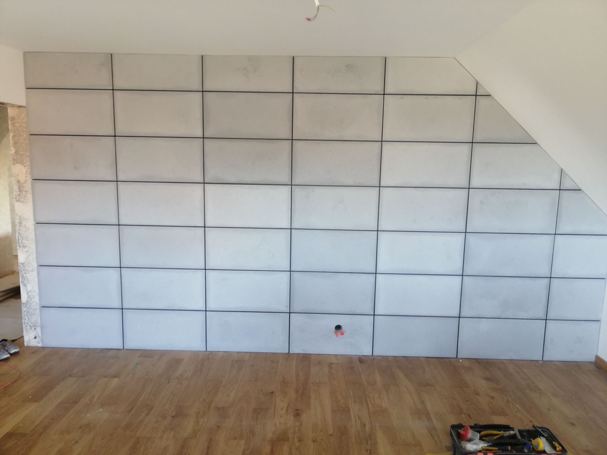 Ściana wykończona panelami imitującymi beton, z widocznym gniazdkiem elektrycznym i narzędziami na podłodze. Poddasze z drewnianą podłogą.