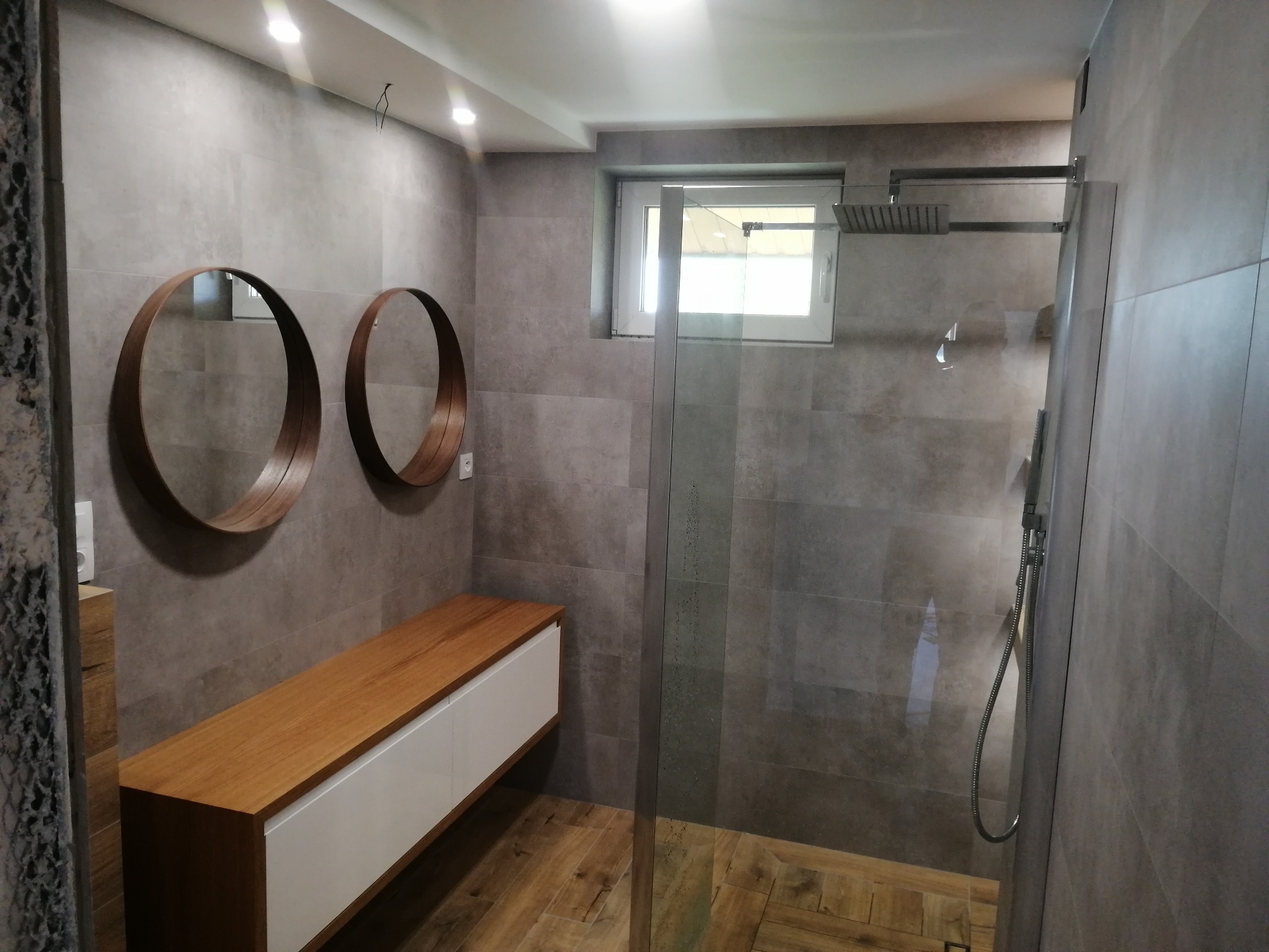 Nowoczesna łazienka z kabiną prysznicową typu walk-in, drewnianą szafką z białymi frontami i okrągłymi lustrami w drewnianych ramach na szarej, płytkowej ścianie.