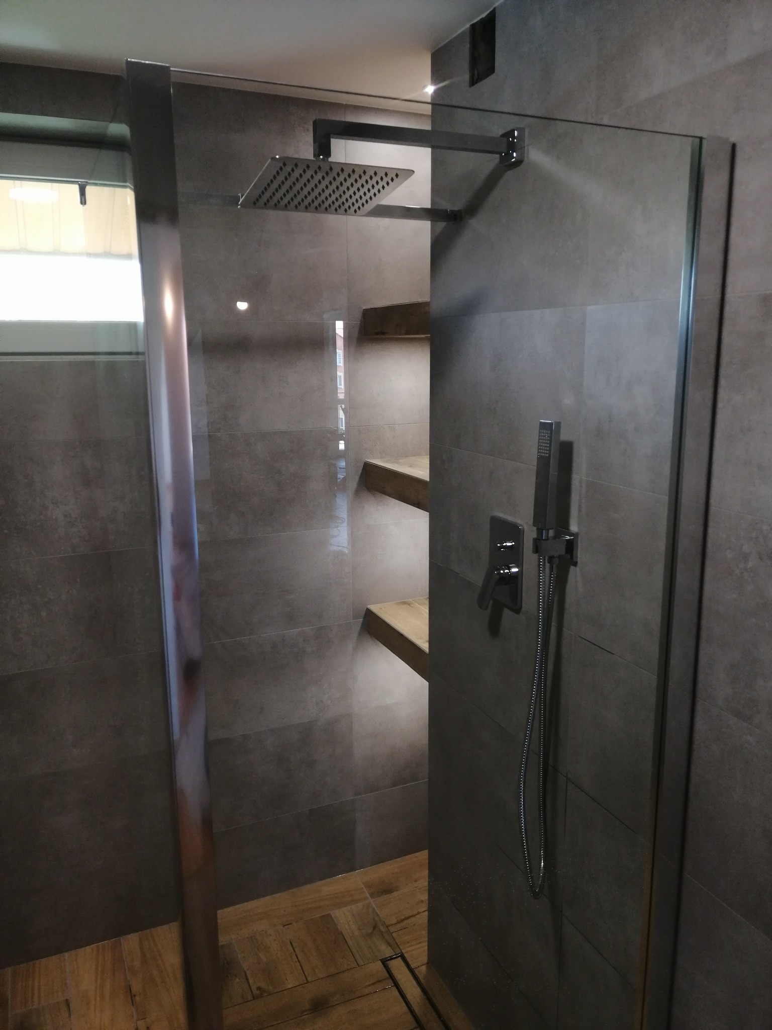 Nowoczesna kabina prysznicowa z deszczownicą i półkami z drewna, wykończona szarą płytką. Minimalistyczny design, widoczne detale armatury i drewniane akcenty.