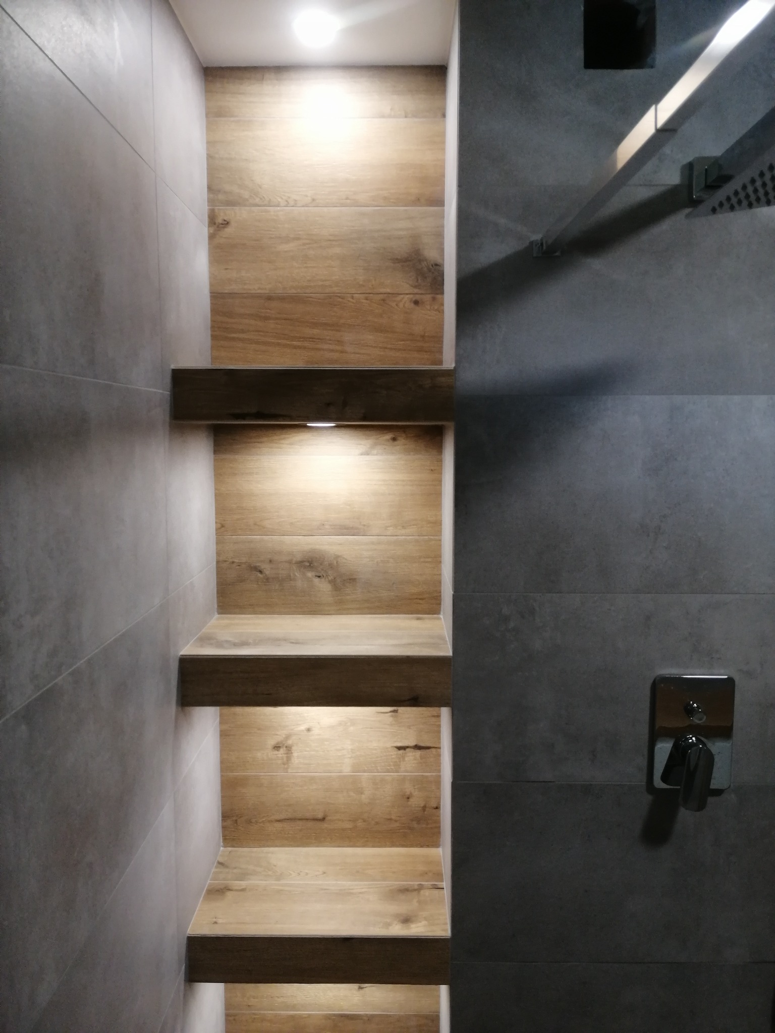 Nowoczesna łazienka w Sośnicy z szarymi płytkami i drewnianymi półkami podświetlonymi LED, chromowana armatura, minimalistyczny design.