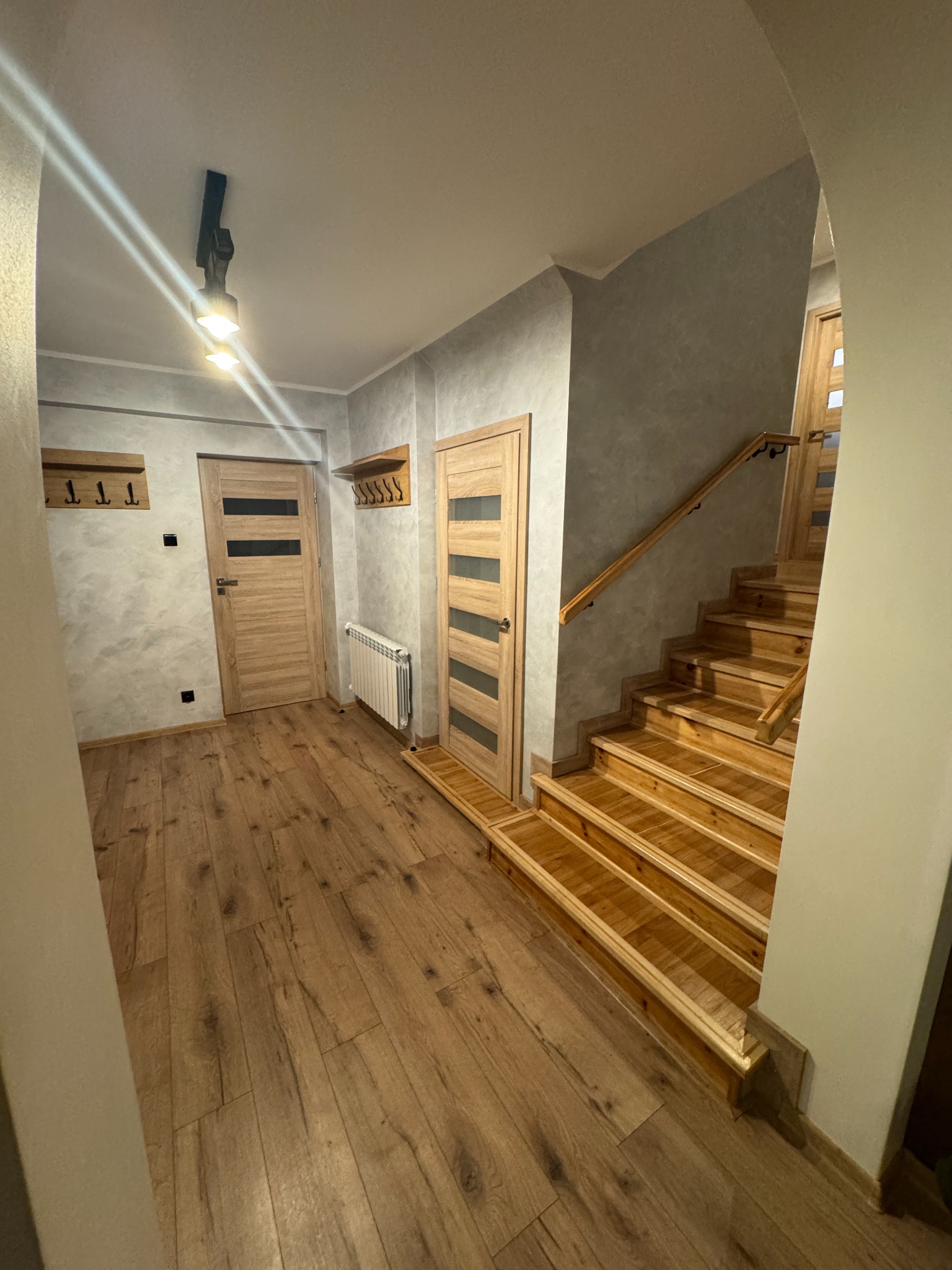 Korytarz z drewnianymi schodami, drzwiami i podłogą. Na ścianie wieszaki i grzejnik. Stonowana kolorystyka, nowoczesny design. Oświetlenie sufitowe.