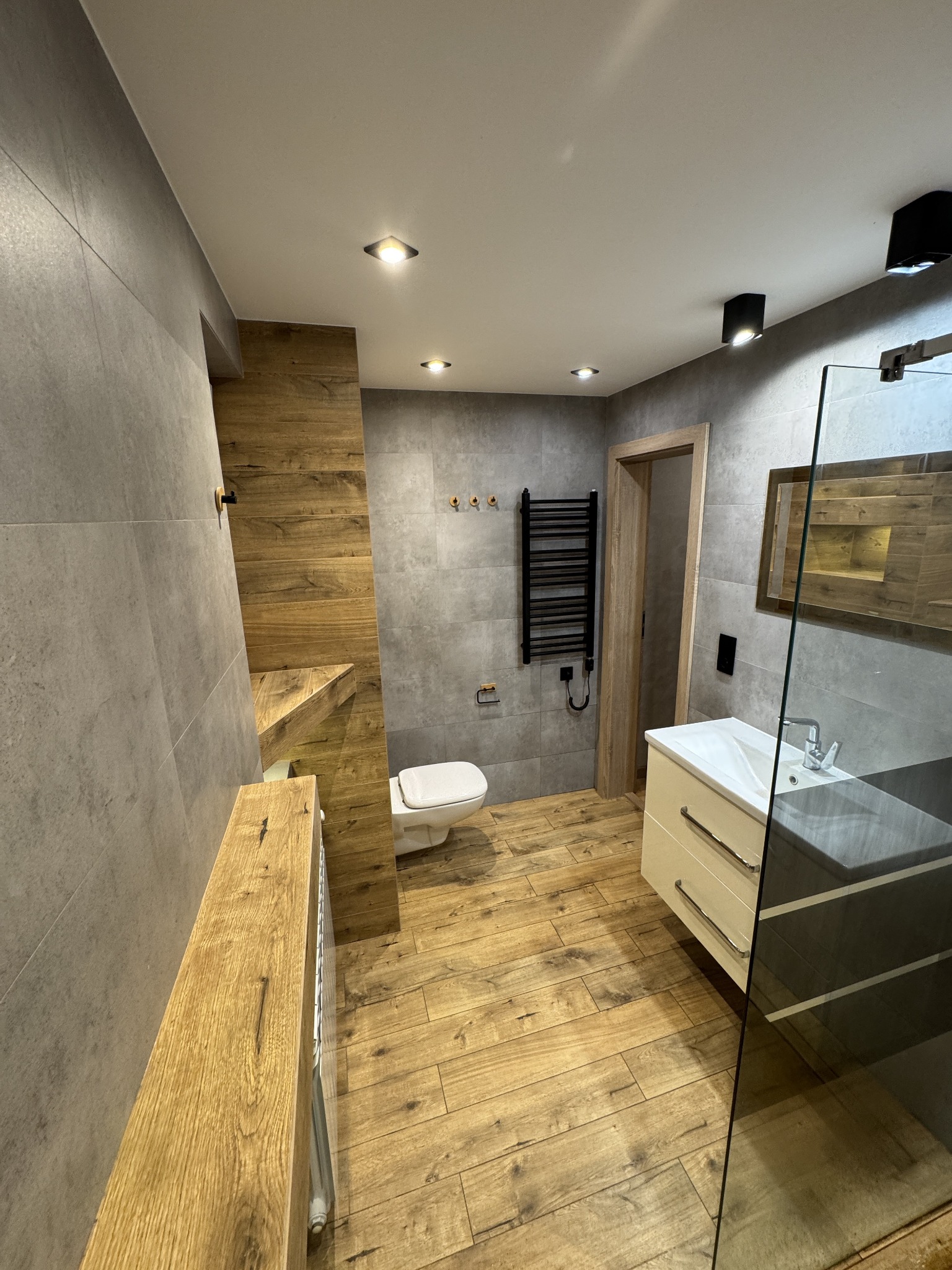 Nowoczesna łazienka z szarymi płytkami, drewnianymi akcentami i czarnym grzejnikiem drabinkowym. Umywalka z szafką i kabina prysznicowa ze szklaną ścianką.