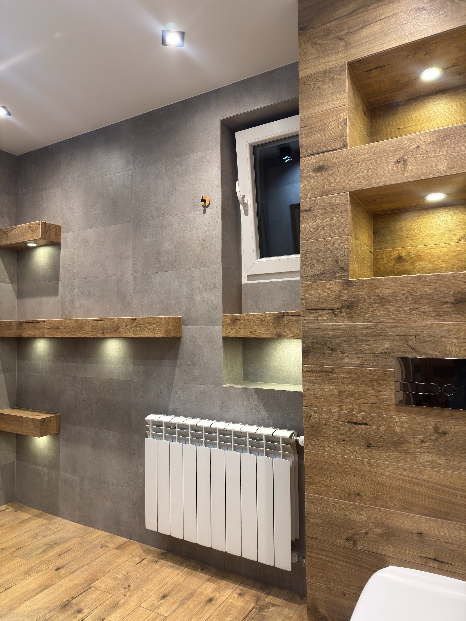 Nowoczesna łazienka z szarymi płytkami, drewnianymi półkami z oświetleniem, białym grzejnikiem i oknem. Elementy drewniane dodają ciepła surowemu wnętrzu.