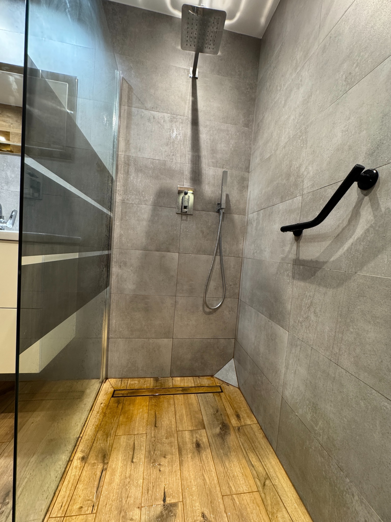 Nowoczesna kabina prysznicowa walk-in z drewnopodobną podłogą i szarą, minimalistyczną armaturą. Widoczny odpływ liniowy i uchwyt dla niepełnosprawnych.