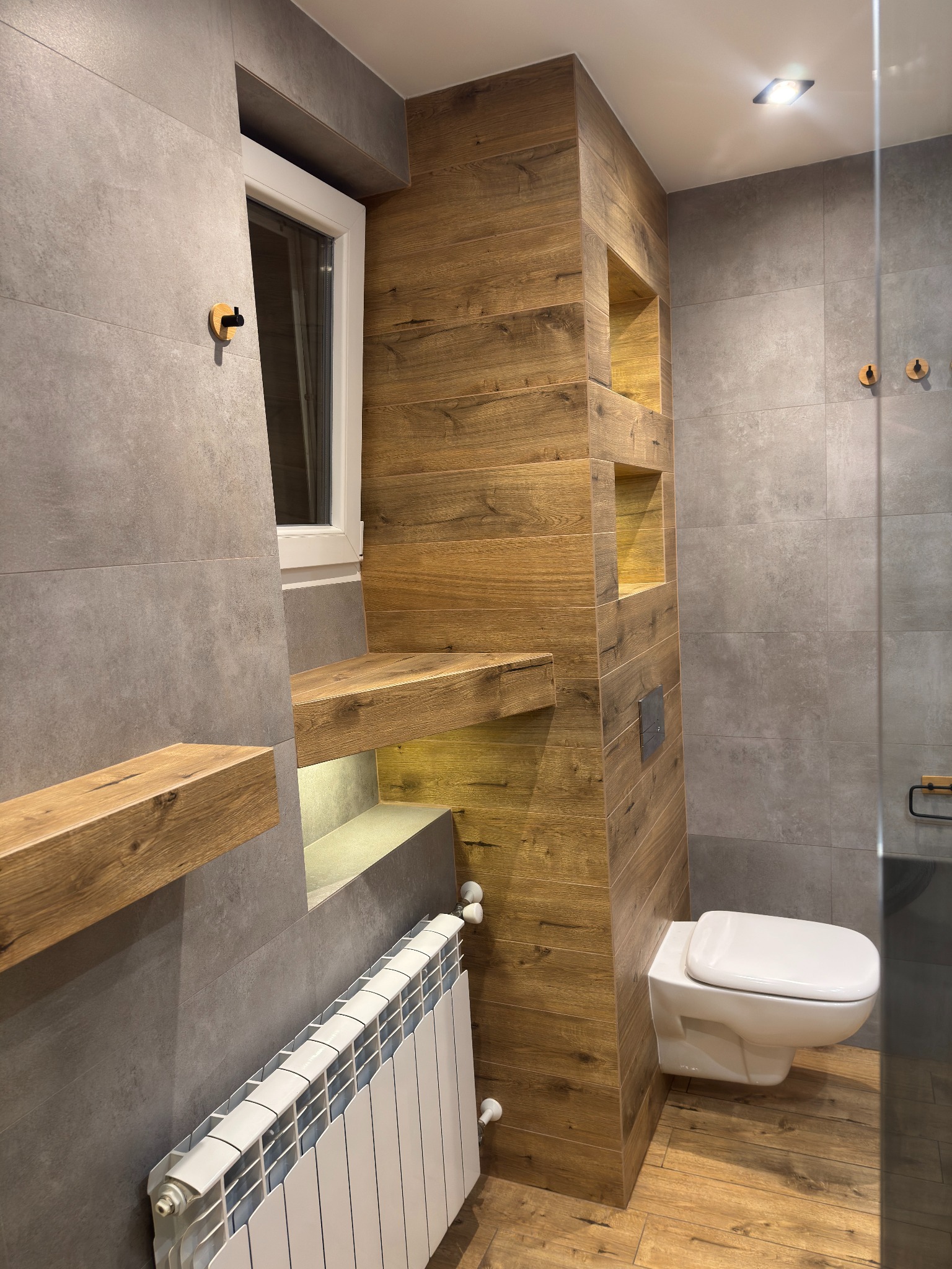 Nowoczesna łazienka z drewnianymi akcentami: półki z podświetleniem, ściana z drewnopodobnych płytek, wisząca toaleta i grzejnik. Szare płytki na ścianach i podłodze.