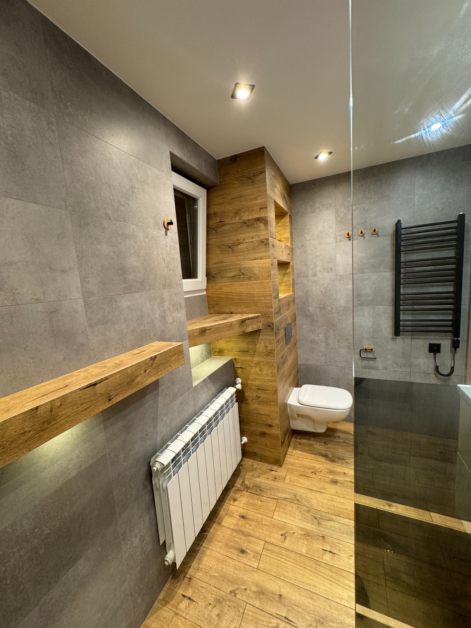 Nowoczesna łazienka z szarymi płytkami, drewnianymi akcentami i grzejnikiem. Widoczne oświetlenie punktowe, toaleta i drewniana półka. Ujęcie z perspektywy.