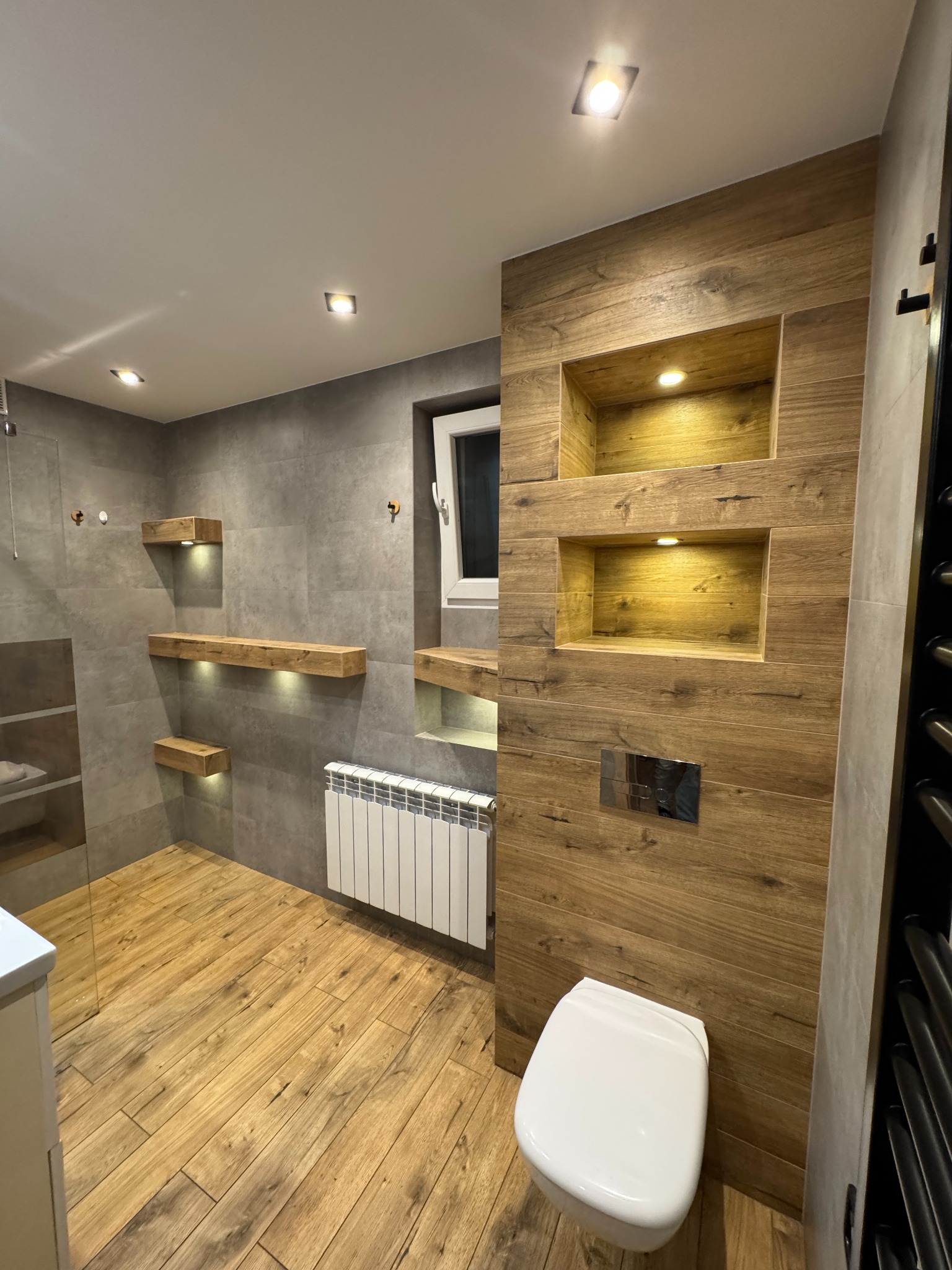 Nowoczesna łazienka z drewnianymi akcentami: półki z podświetleniem, grzejnik, toaleta. Szare płytki i drewnopodobna podłoga tworzą ciepły klimat.