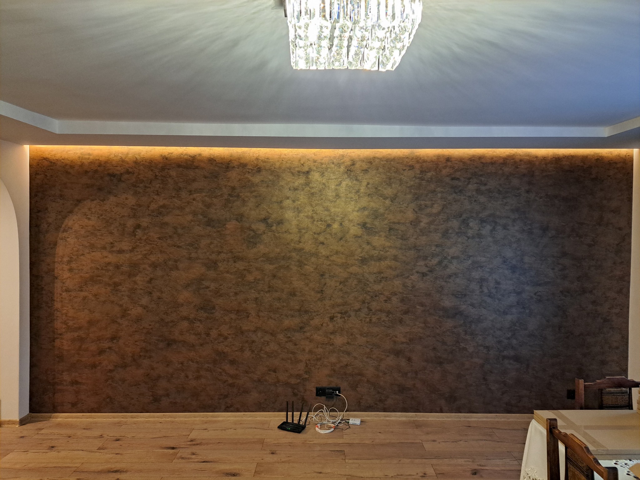 Ściana z efektem dekoracyjnym w odcieniach brązu i złota, oświetlona listwą LED. Na suficie kryształowy żyrandol. Parkiet na podłodze. Widoczny router i kable.