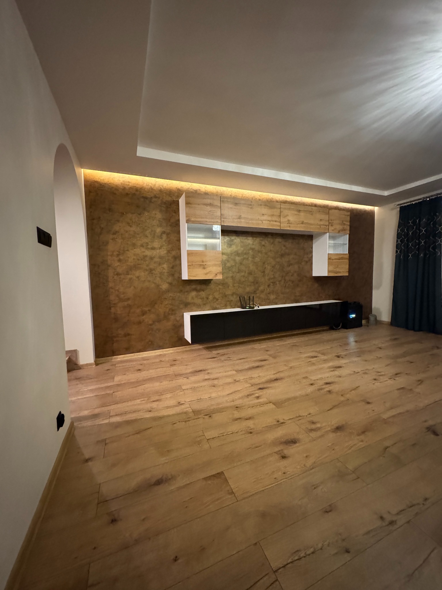 Nowoczesny salon z drewnianą podłogą, ścianą w odcieniach brązu i zabudową meblową z oświetleniem LED. Widoczny łuk wejściowy i zasłony w oknie.