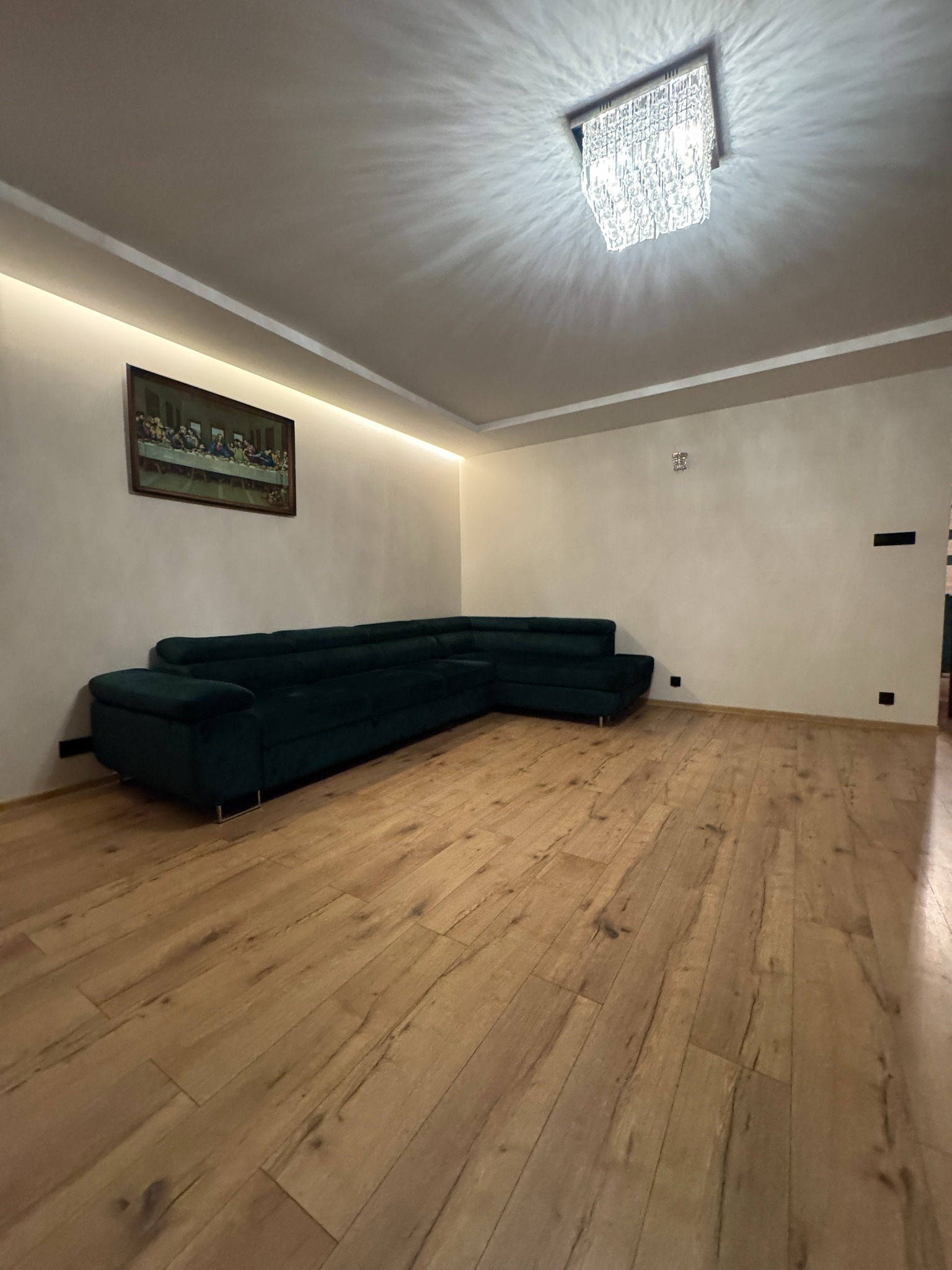 Salon z parkietem, narożnikiem w odcieniach zieleni i obrazem na ścianie. Sufit zdobi kryształowy żyrandol, a oświetlenie LED podkreśla wystrój.