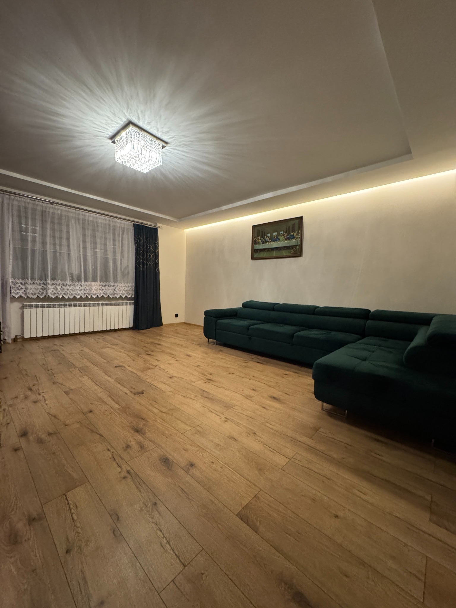 Salon z drewnianą podłogą, zieloną sofą narożną i kryształowym żyrandolem. Ściany w ciepłym odcieniu, obraz na ścianie. Okno z firanką i zasłoną.