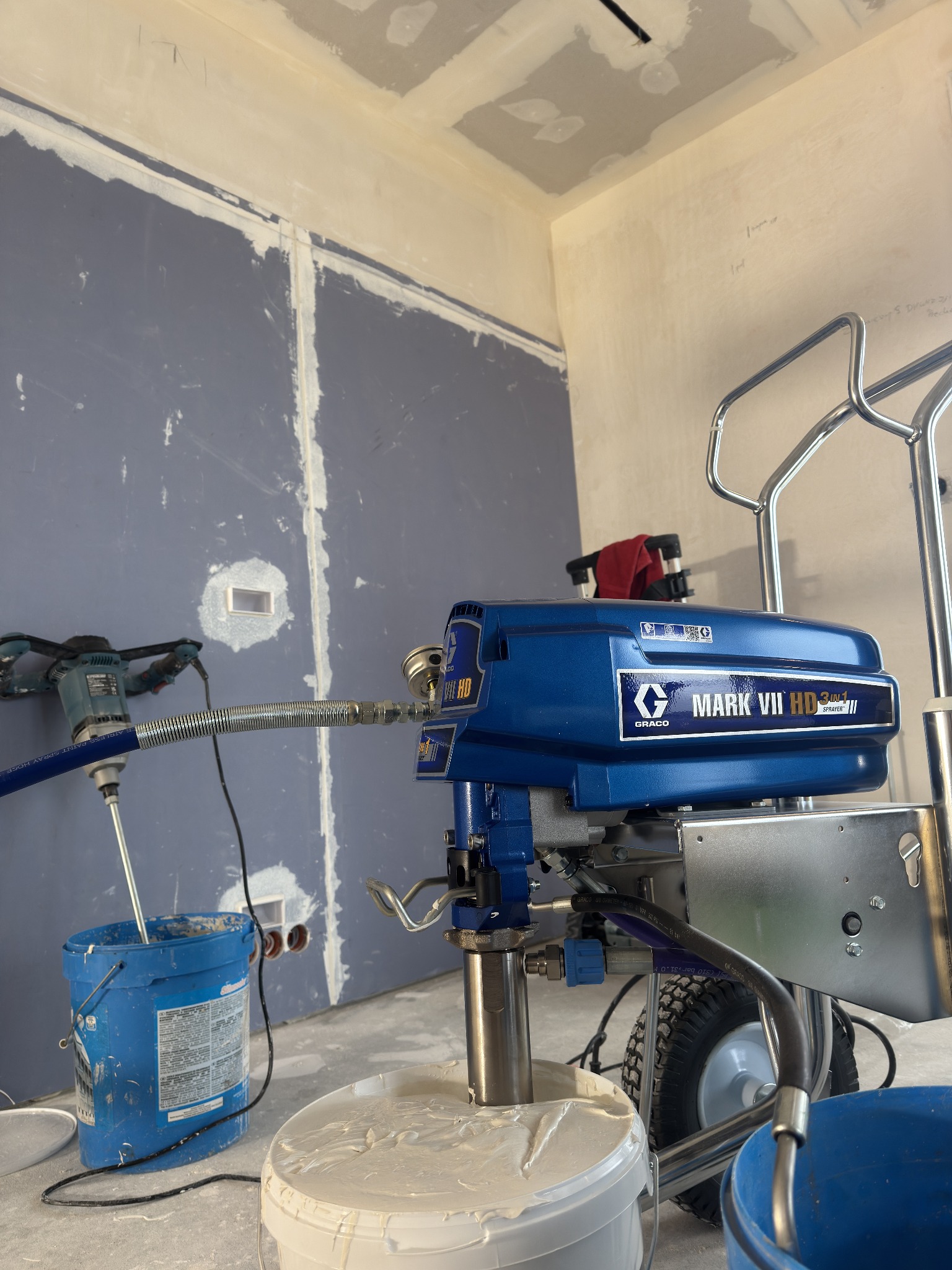 Agregat malarski Graco Mark VII HD z mieszadłem w wiadrze, przygotowany do pracy w pomieszczeniu z płytami gipsowo-kartonowymi przed malowaniem. Widoczne ślady szpachlowania.