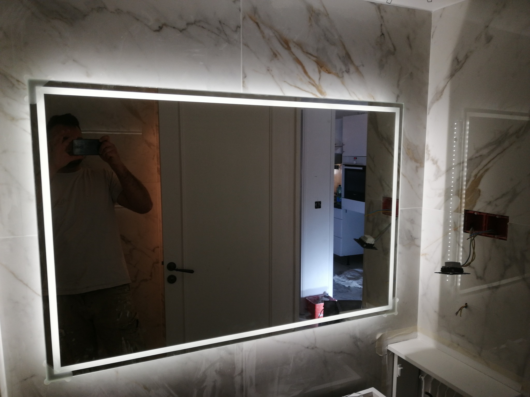 Nowoczesne lustro LED w łazience z marmurowymi płytkami. Widoczne odbicie osoby robiącej zdjęcie. Elementy instalacji elektrycznej na ścianie.
