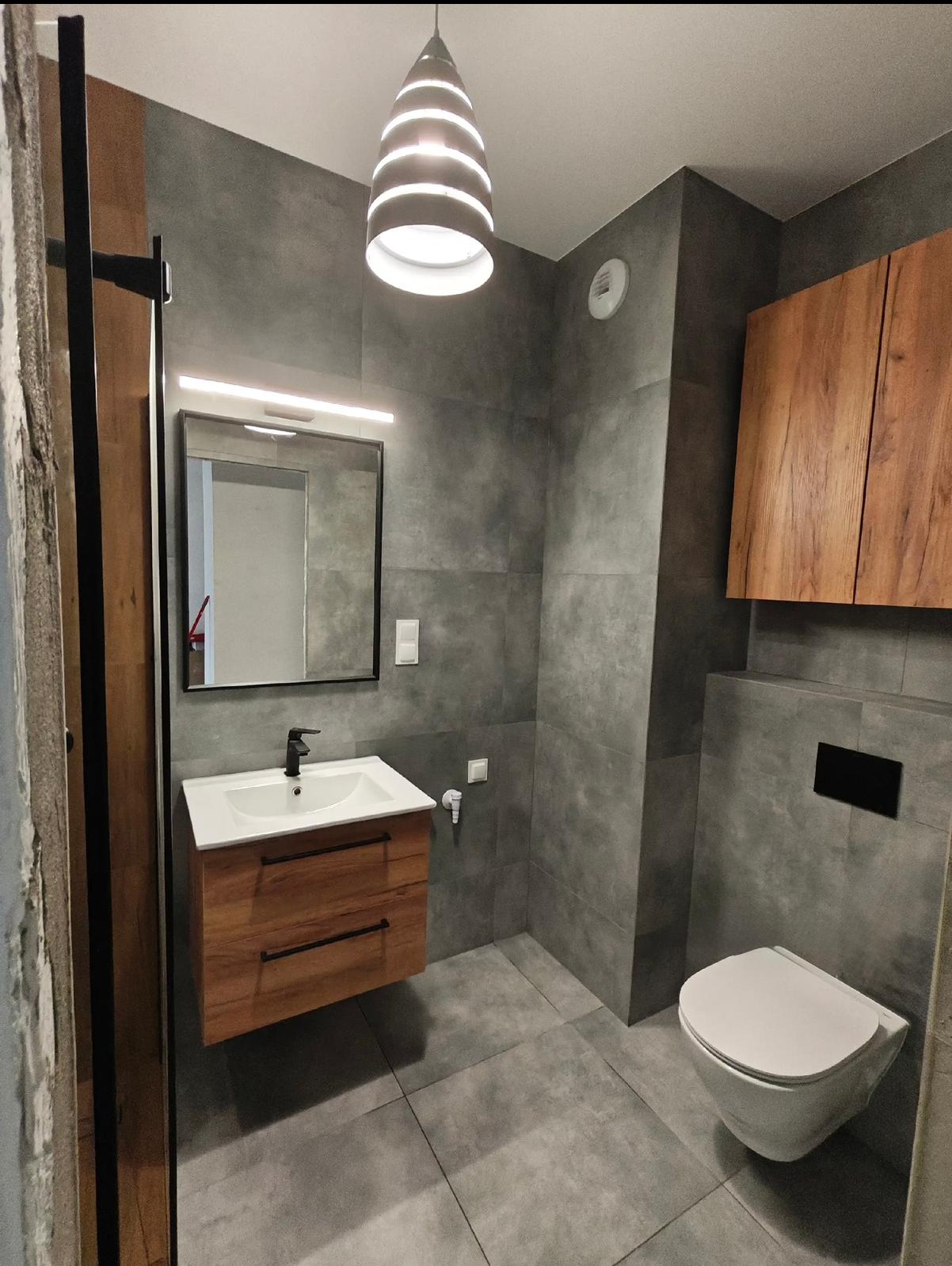 Nowoczesna łazienka w szarościach z drewnianymi akcentami: szafka pod umywalką i wisząca szafka. Uwagę przykuwa designerska lampa sufitowa.