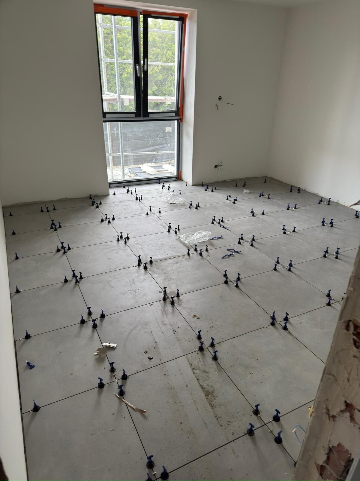 Układanie szarych płytek podłogowych z użyciem niebieskich klipsów poziomujących w jasnym pokoju z oknem. Widoczne fugi i fragment drabiny.