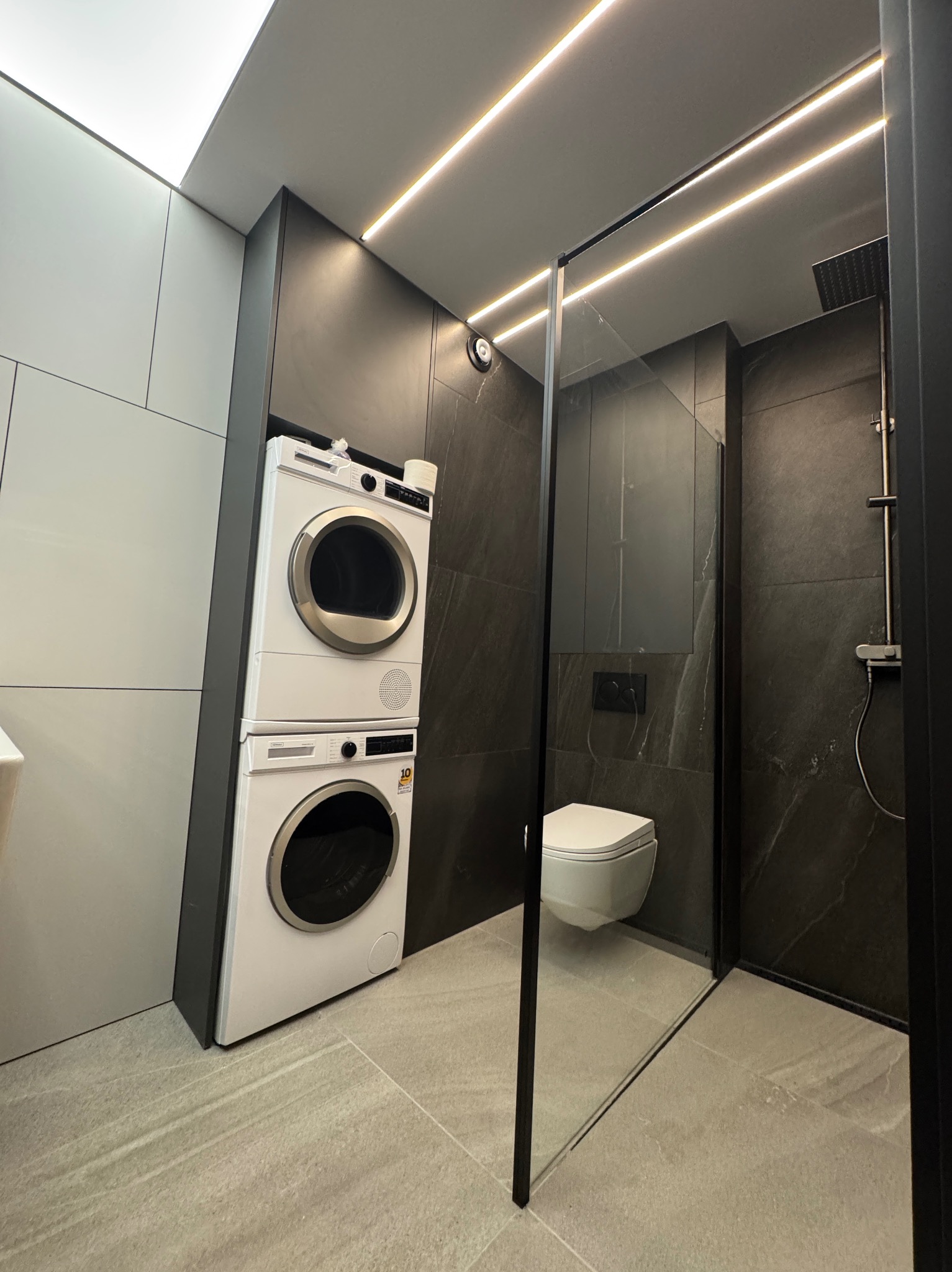 Nowoczesna łazienka z kabiną prysznicową typu walk-in, toaletą podwieszaną i pralko-suszarką w zabudowie. Minimalistyczny design, szare płytki i oświetlenie LED.