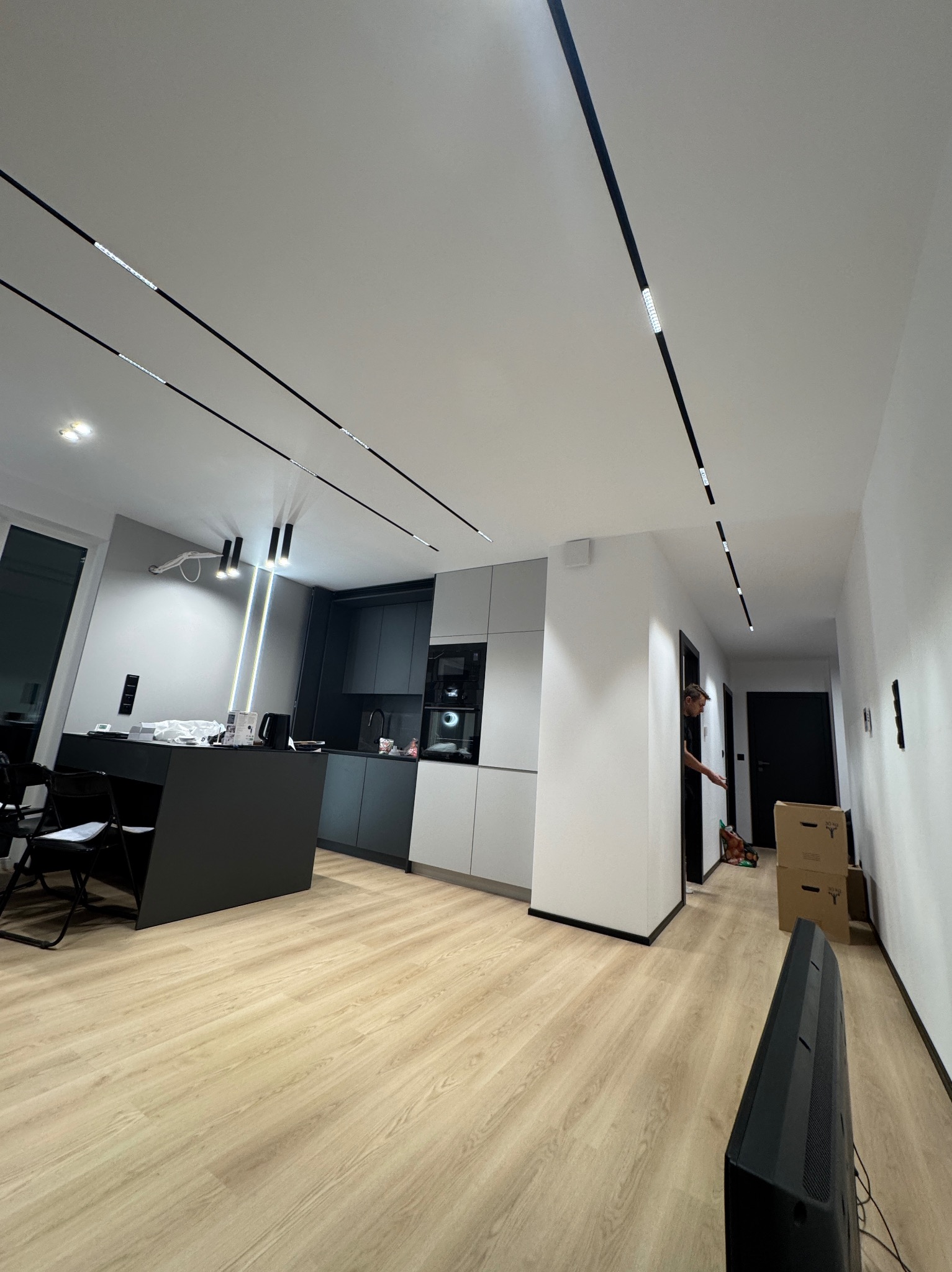 Nowoczesne wnętrze: czarno-szara kuchnia z wyspą, minimalistyczne oświetlenie liniowe na suficie, jasna podłoga. Osoba w tle przy drzwiach.