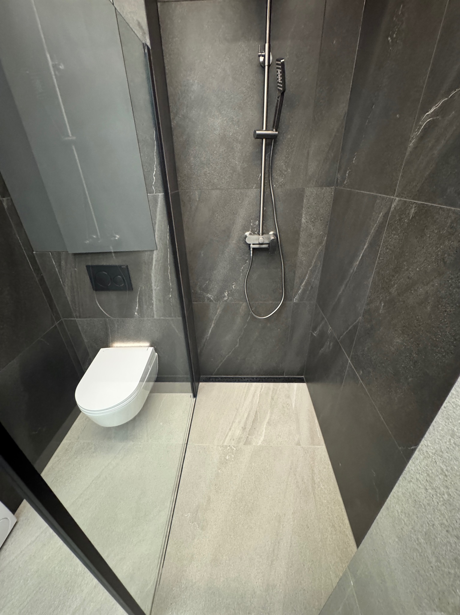 Nowoczesna łazienka z kabiną prysznicową typu walk-in, wiszącą toaletą i szarymi płytkami imitującymi kamień. Minimalistyczny design z czarnymi akcentami.