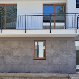 Tomasz Kądziołka usługi budowlane i ogrodnicze - Elewacja domu z szarymi płytami imitującymi kamień, widoczne okna z drewnianą ramą i balkon z czarną balustradą. Częściowo widoczny teren budowy.