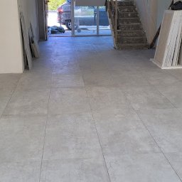 Tomasz Kądziołka usługi budowlane i ogrodnicze - Ułożona, jasnoszara podłoga z płytek w przestronnym wnętrzu. Widoczne przygotowane płyty gipsowe i schody w tle. Częściowo wykończone ściany z widocznymi puszkami elektrycznymi.