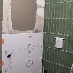 Tomasz Kądziołka usługi budowlane i ogrodnicze - Wnętrze łazienki w trakcie prac. Na ścianach białe i zielone płytki, widoczne krzyżyki dystansowe. Na podłodze wiadro z zaprawą i worek kleju.