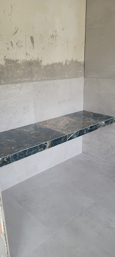 Półka z ciemnego marmuru z wyraźnym żyłkowaniem, wkomponowana w szarą, wykafelkowaną wnękę. Ściana częściowo surowa, z widocznymi śladami prac wykończeniowych.