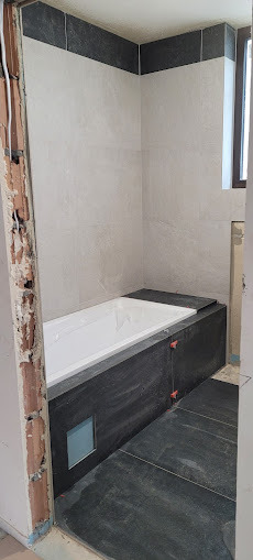 Wanna obudowana ciemnymi płytkami, jasne płytki na ścianach, widoczne krzyżyki glazurnicze, surowy fragment ściany. Ukończony etap prac w łazience.