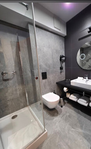 Nowoczesna łazienka z kabiną prysznicową typu walk-in, umywalką na blacie i toaletą. Szare płytki imitujące marmur na ścianach i podłodze. Minimalistyczne oświetlenie.