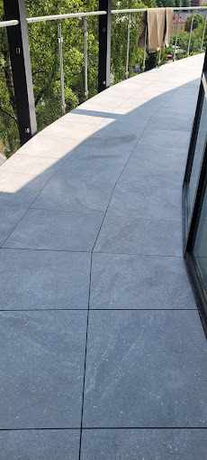 Wykończony balkon z szarymi płytkami, widok z góry. Balustrada ze stali nierdzewnej i czarnych elementów. Częściowo zacieniona powierzchnia. Suszarka na pranie w tle.