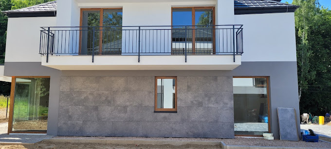 Elewacja domu z szarymi płytami imitującymi kamień, widoczne okna z drewnianą ramą i balkon z czarną balustradą. Częściowo widoczny teren budowy.