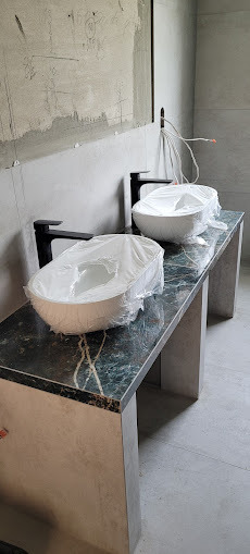 Dwie białe umywalki nablatowe, jeszcze w folii ochronnej, stoją na blacie z zielonego marmuru w surowej łazience z częściowo położonymi płytkami.