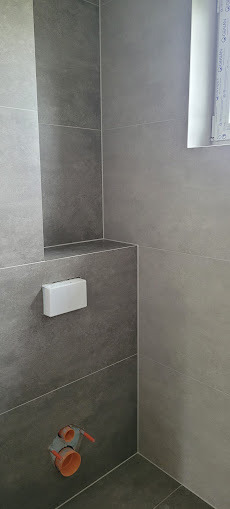 Szara, nowoczesna łazienka z ukrytą spłuczką i widocznymi rurami kanalizacyjnymi. Minimalistyczny design, płytki imitujące beton, okno w tle.