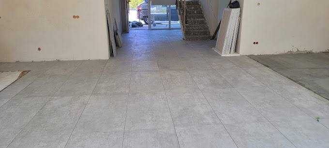 Ułożona, jasnoszara podłoga z płytek w przestronnym wnętrzu. Widoczne przygotowane płyty gipsowe i schody w tle. Częściowo wykończone ściany z widocznymi puszkami elektrycznymi.