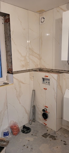 Białe płytki imitujące marmur na ścianach łazienki w trakcie montażu. Widoczne elementy instalacji sanitarnej i narzędzia glazurnicze na podłodze. Częściowo wykończone wnętrze.